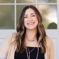 Smiling woman with long wavy brown hair wearing a tan cardigan and layered necklaces.